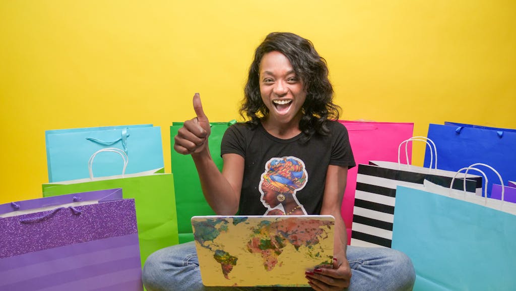 Cheerful woman using a laptop surrounded by colorful shopping bags, showing a thumbs up sign.